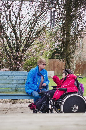 A person in a blue outdoor jacket is sat on a bench outside, talking to a person in a red coat who is in a wheelchair