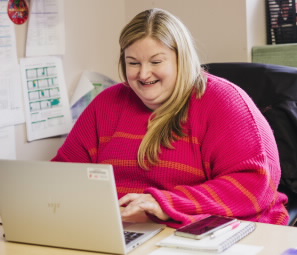 A woman is sat at a desk using a laptop. She has blonde hair and is wearing a pink jumper.