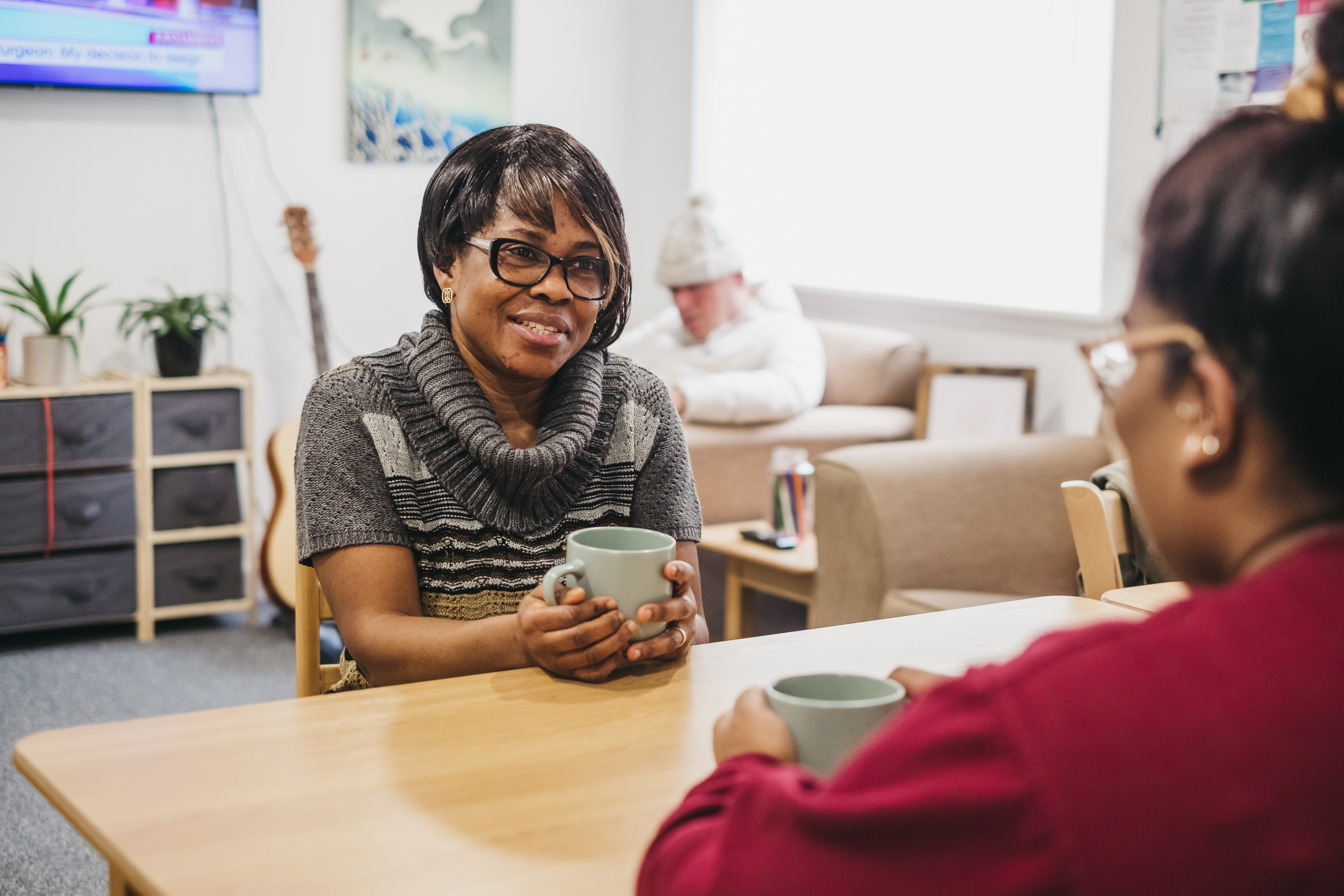 A lady sitting at a wooden table speaking to another lady, both enjoying a hot drink